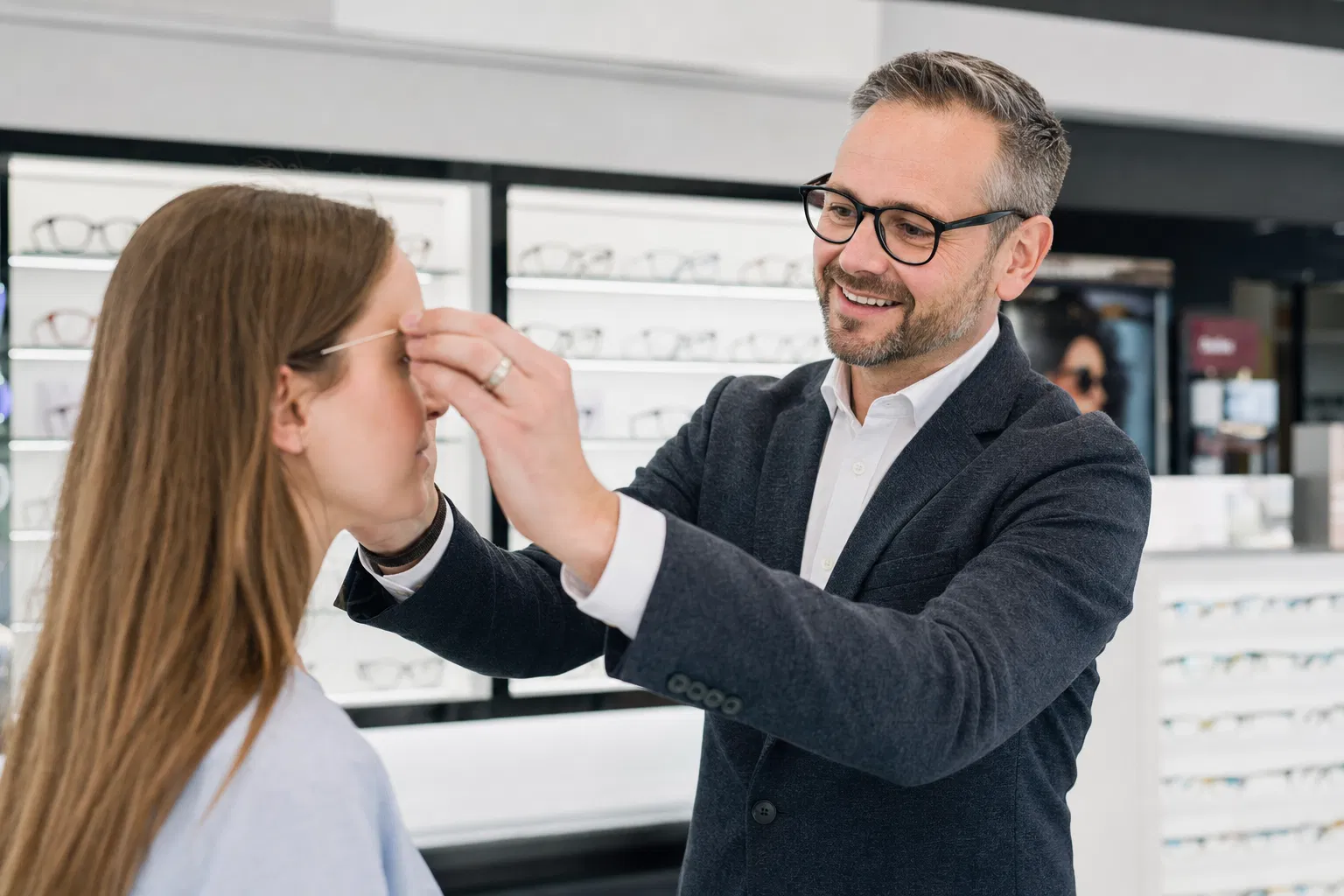 Augenoptiker passt einer Kundin im Geschäft eine Brille an, im Hintergrund beleuchtete Regale mit Brillenfassungen