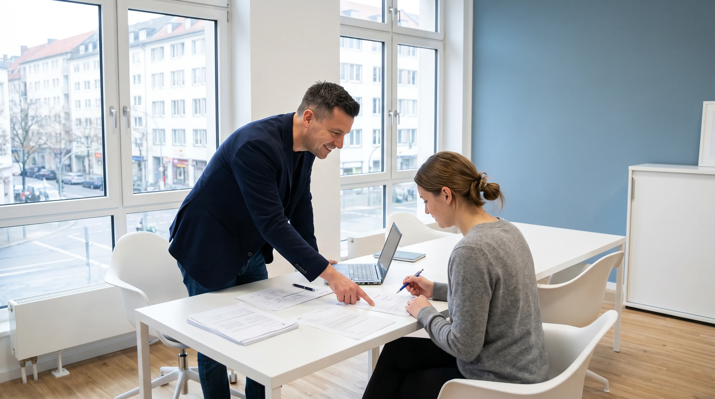 Berater beugt sich in einem hellen Büro über einen Tisch und zeigt einer Frau am Laptop etwas in ausgedruckten Unterlagen