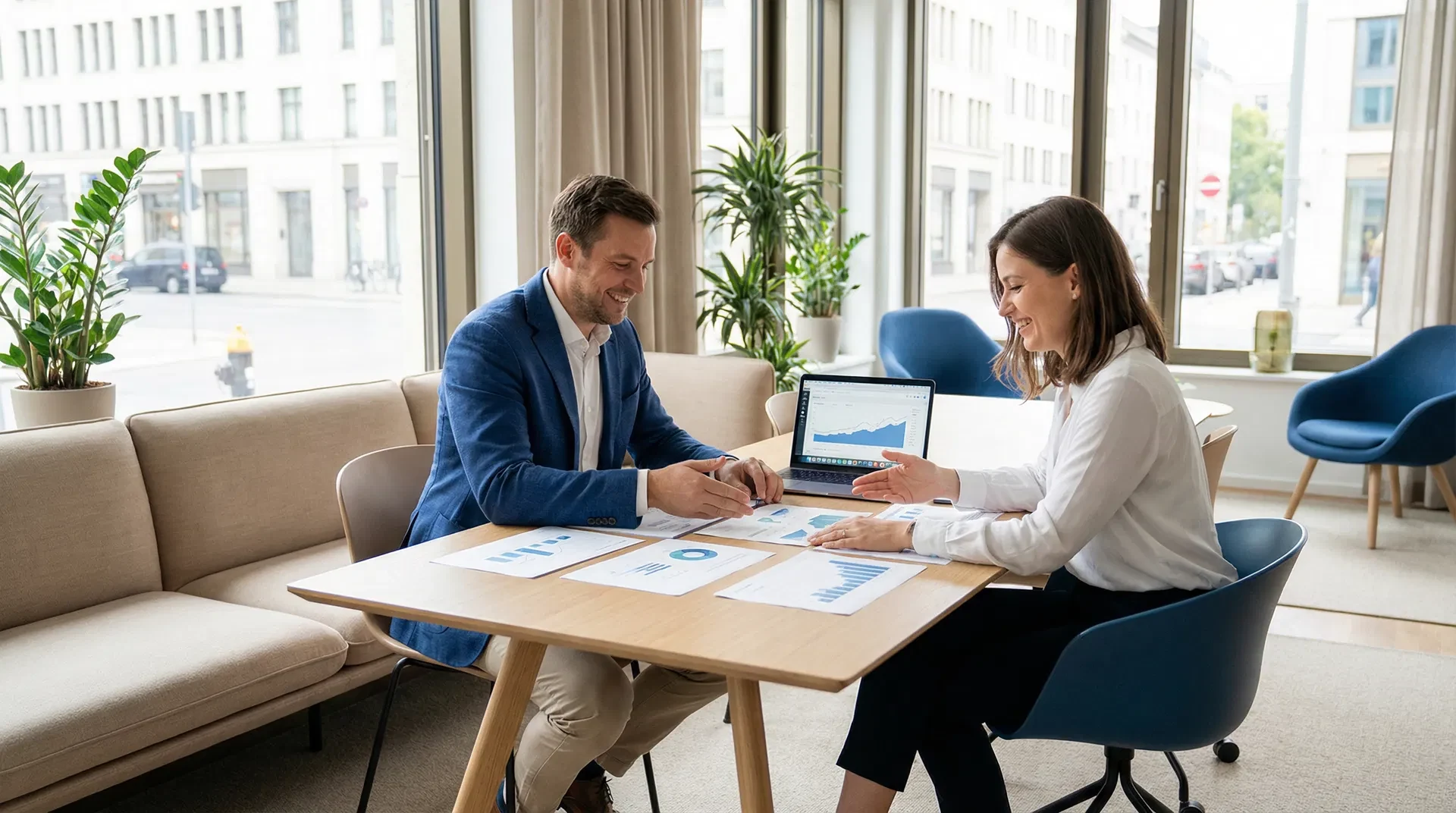 Mann im blauen Sakko und Frau in weißer Bluse besprechen lächelnd Unterlagen an einem Tisch, daneben ein Laptop mit Diagramm