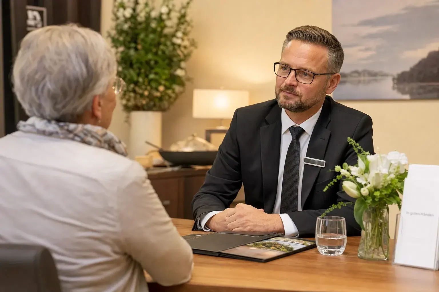 Bestatter im dunklen Anzug hört einer älteren Frau im Beratungsgespräch an einem Tisch mit Blumen zu