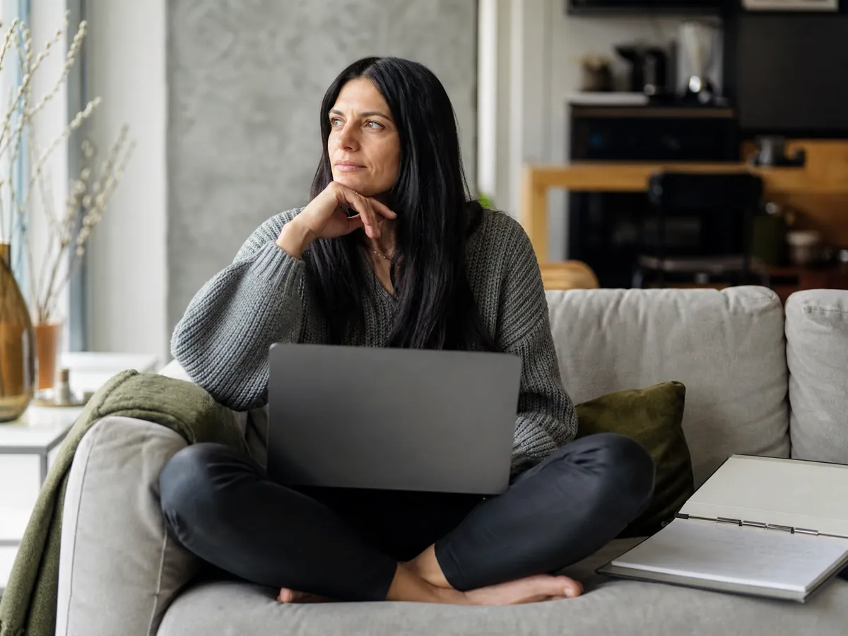 Gründerin wägt auf dem Sofa zwischen Businessplan-Software am Laptop und ausgedruckter Vorlage ab