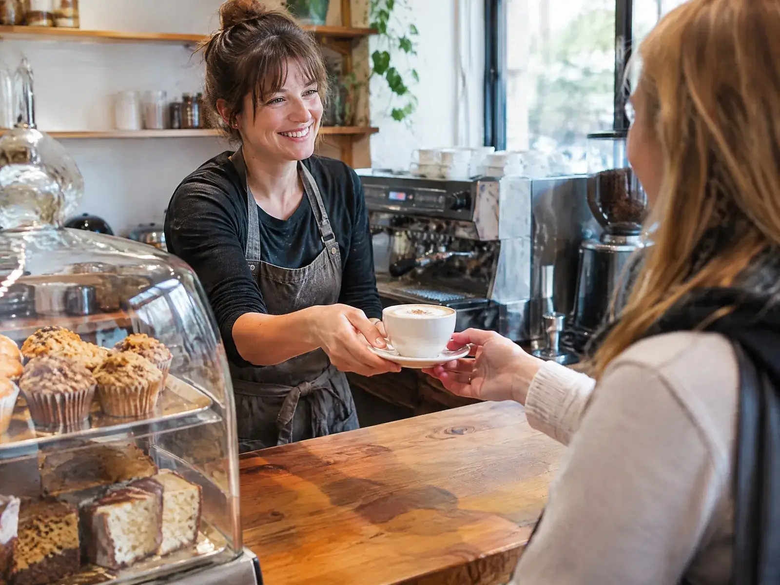 Café eröffnen: Inhaberin reicht einer Kundin an der Holztheke einen Cappuccino, daneben die Kuchenvitrine