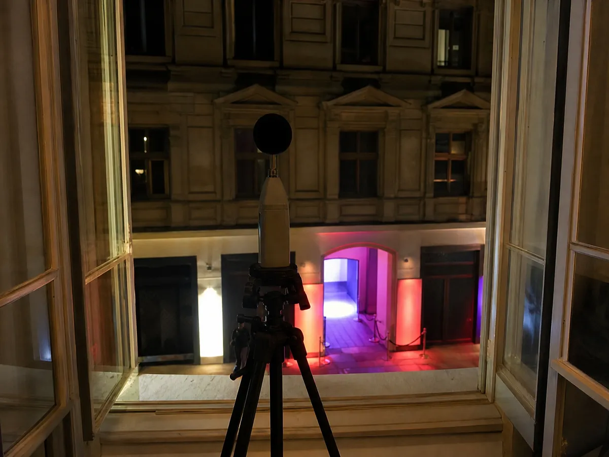 Schallpegelmessgerät am Fenster einer Nachbarwohnung misst nachts den Lärm eines Clubs gegenüber