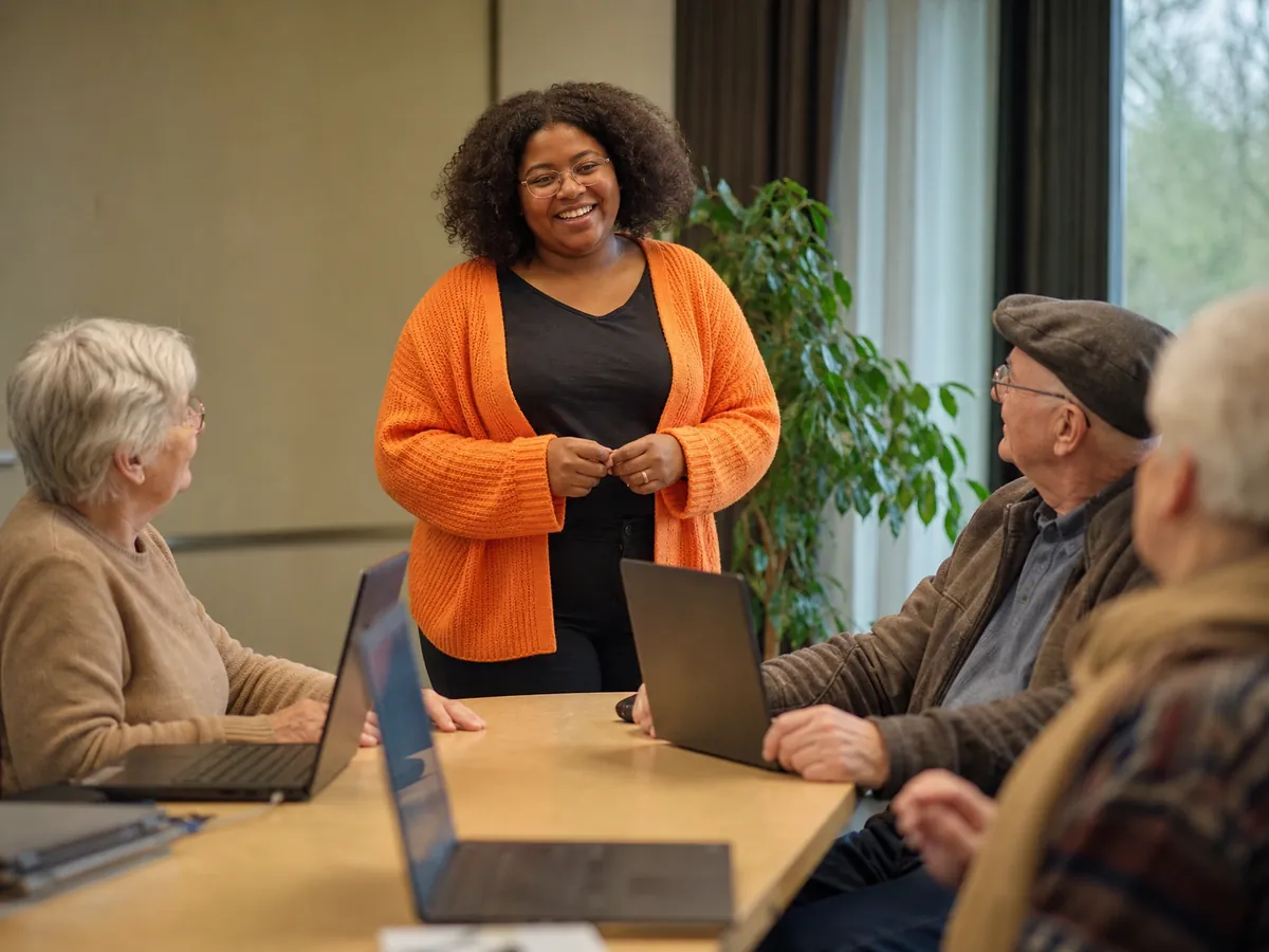 Kursleiterin begrüßt im Gemeinderaum eine Gruppe Senioren zum Computerkurs am Laptop