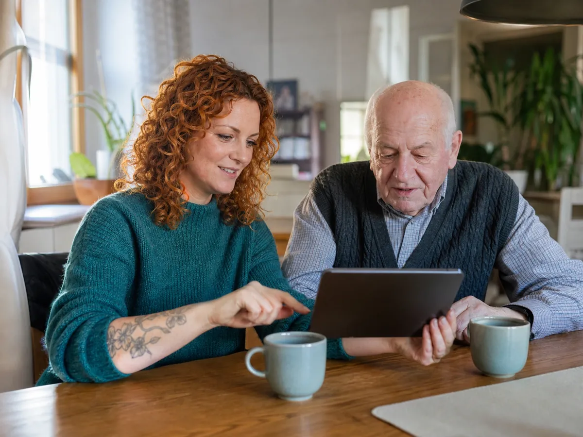 Kursleiterin erklärt einem Senior bei Einzelunterricht zu Hause, wie er das Tablet bedient