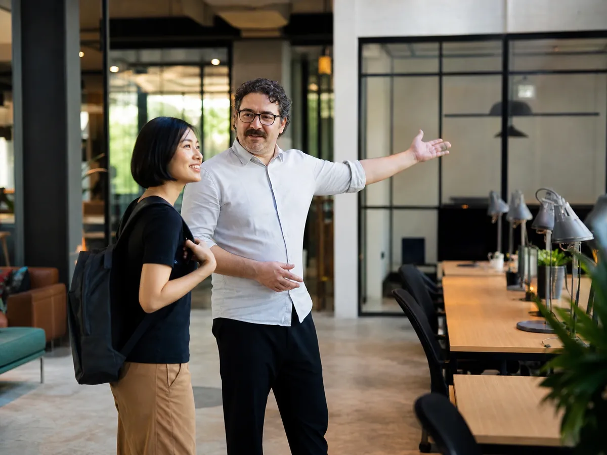 Betreiber zeigt einer neuen Nutzerin im Coworking Space feste Schreibtische und Glasbüros