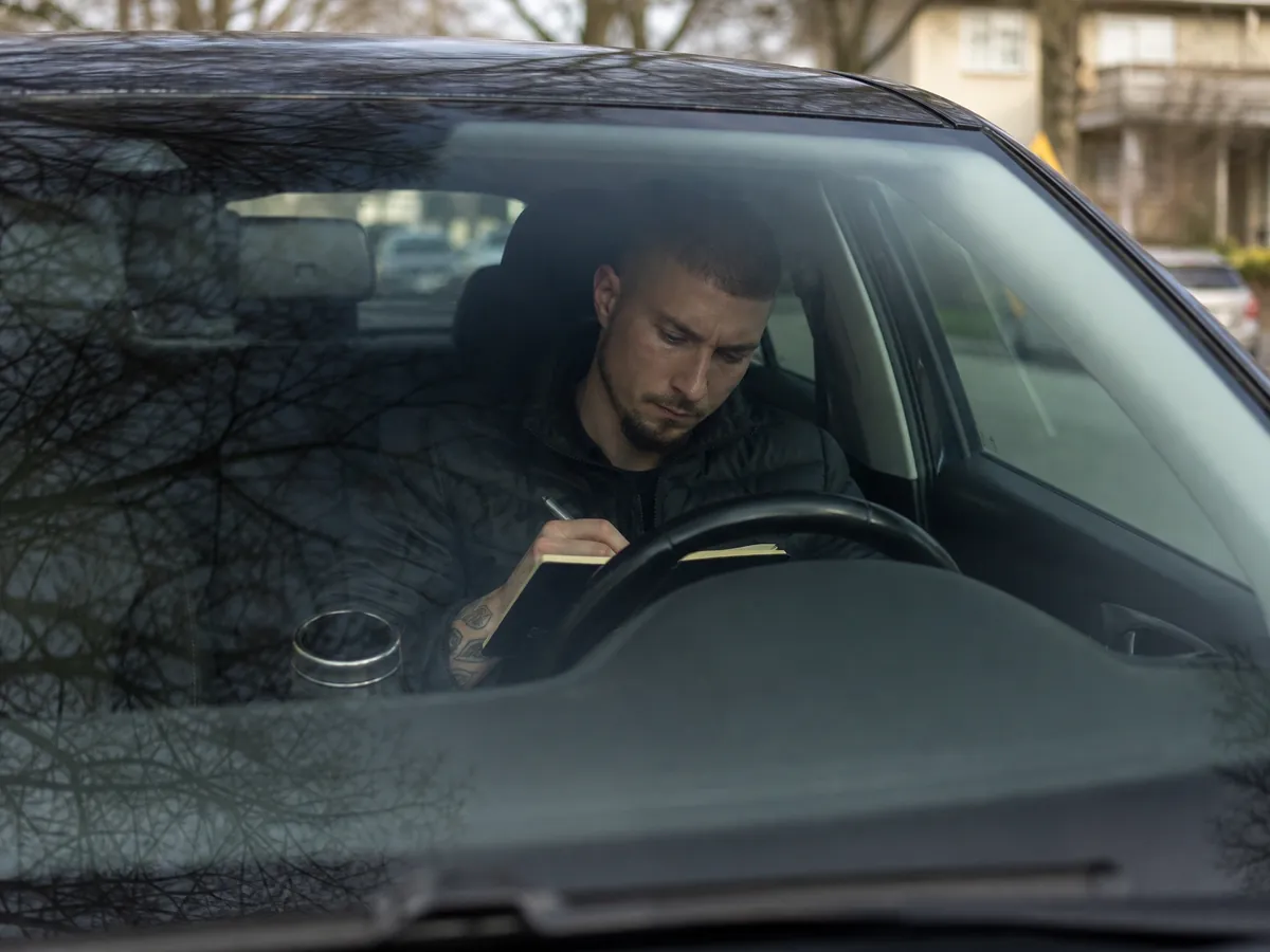 Detektiv beobachtet aus dem Auto eine Wohnstraße und notiert Uhrzeiten in sein Notizbuch