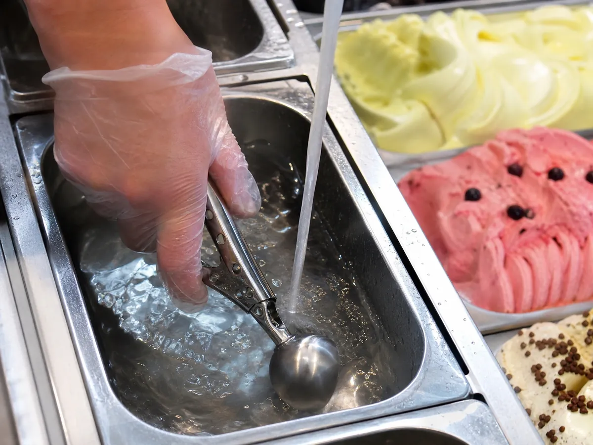 Hand stellt an der Eistheke einen Portionierer in fließendes Wasser, wie es die Hygiene verlangt