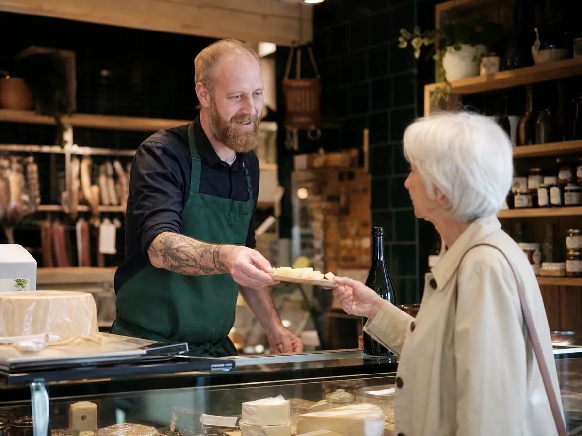 Inhaber eines Feinkostladens lässt eine Kundin an der Theke ein Stück Käse verkosten