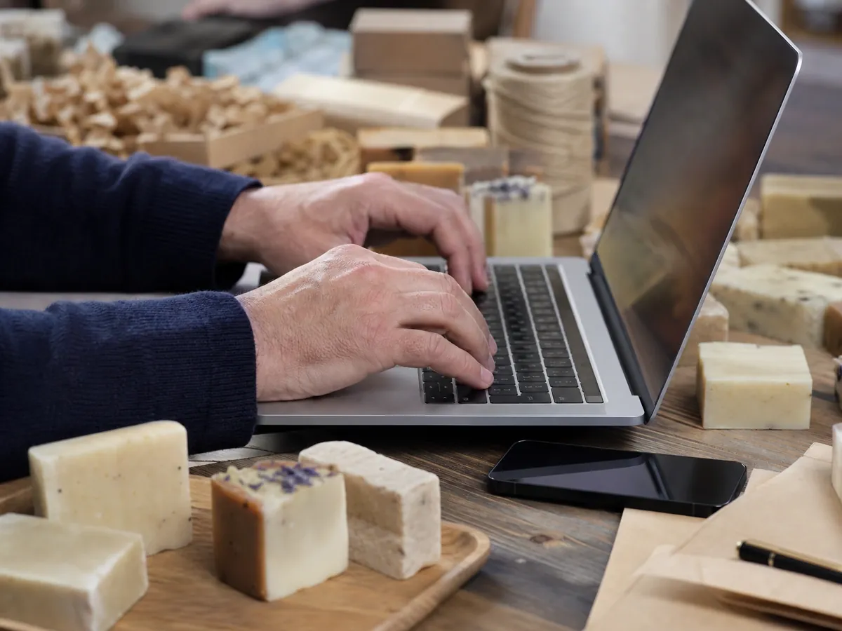 Hände prüfen am Laptop zwischen Seifenmustern, ob Domain und Profilnamen für die Firma frei sind