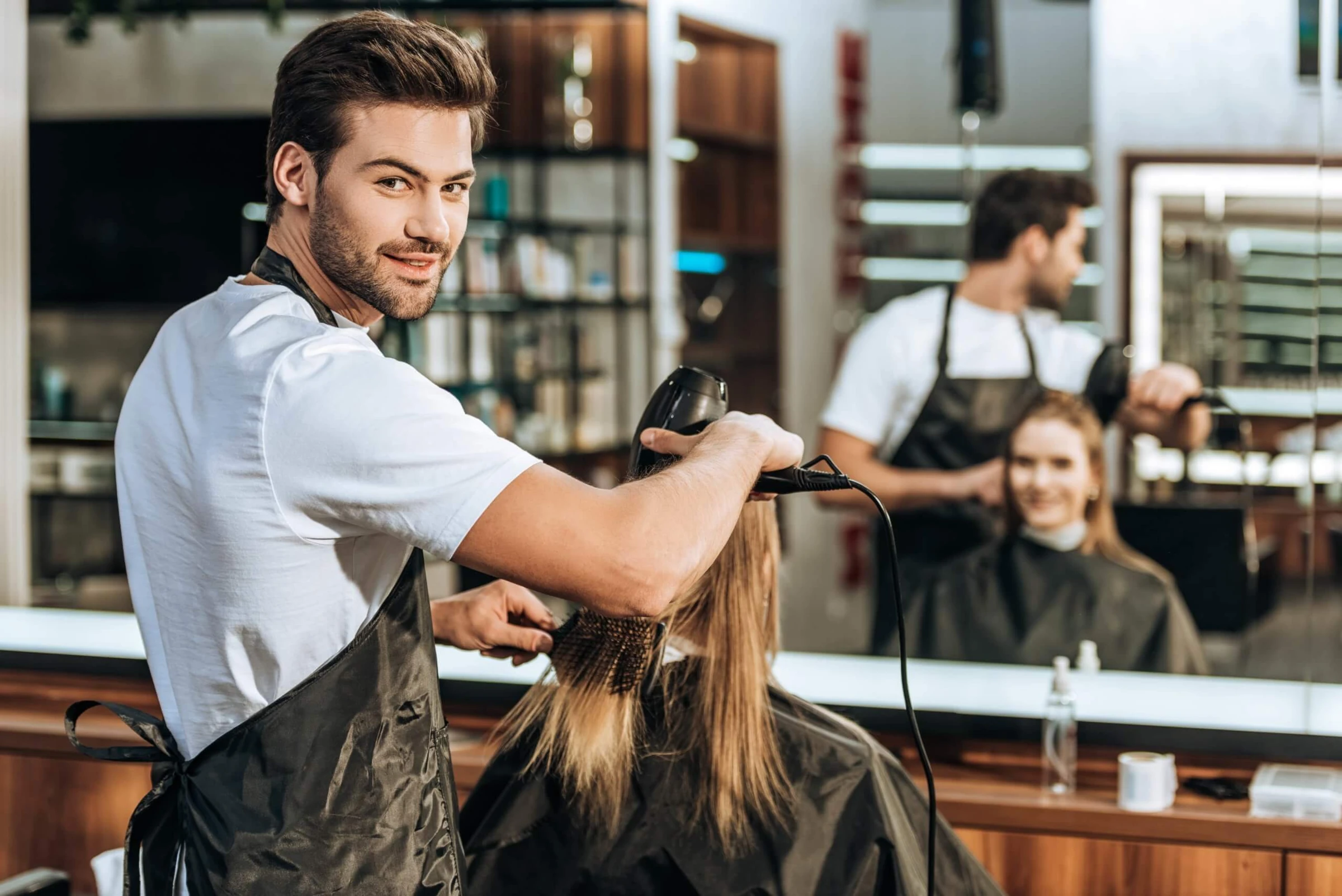 Junger Friseur mit Bart föhnt einer Kundin die Haare und lächelt in die Kamera, im Hintergrund ein Kollege an einem zweiten Platz