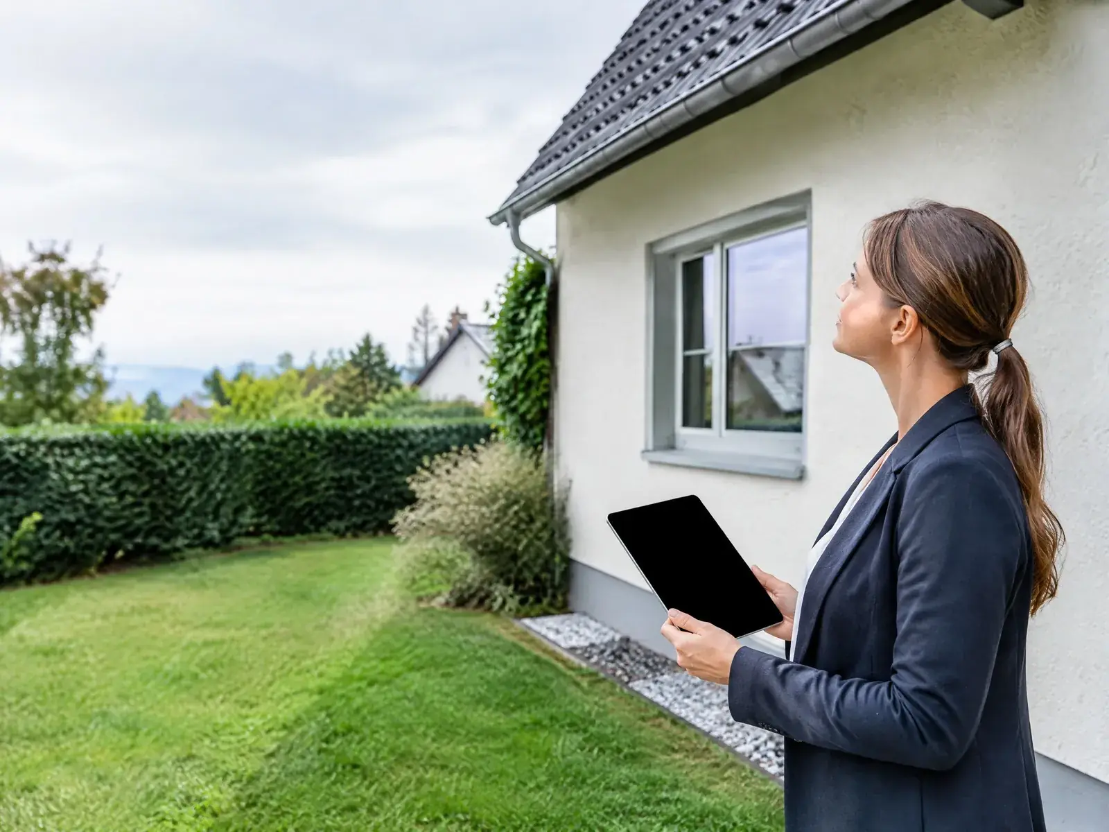 Immobiliengutachterin mit Tablet begutachtet ein Einfamilienhaus von außen