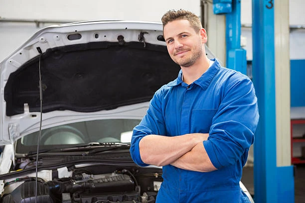 Mann in blauer Arbeitskleidung steht mit verschränkten Armen vor einem Auto mit geöffneter Motorhaube in einer Werkstatt