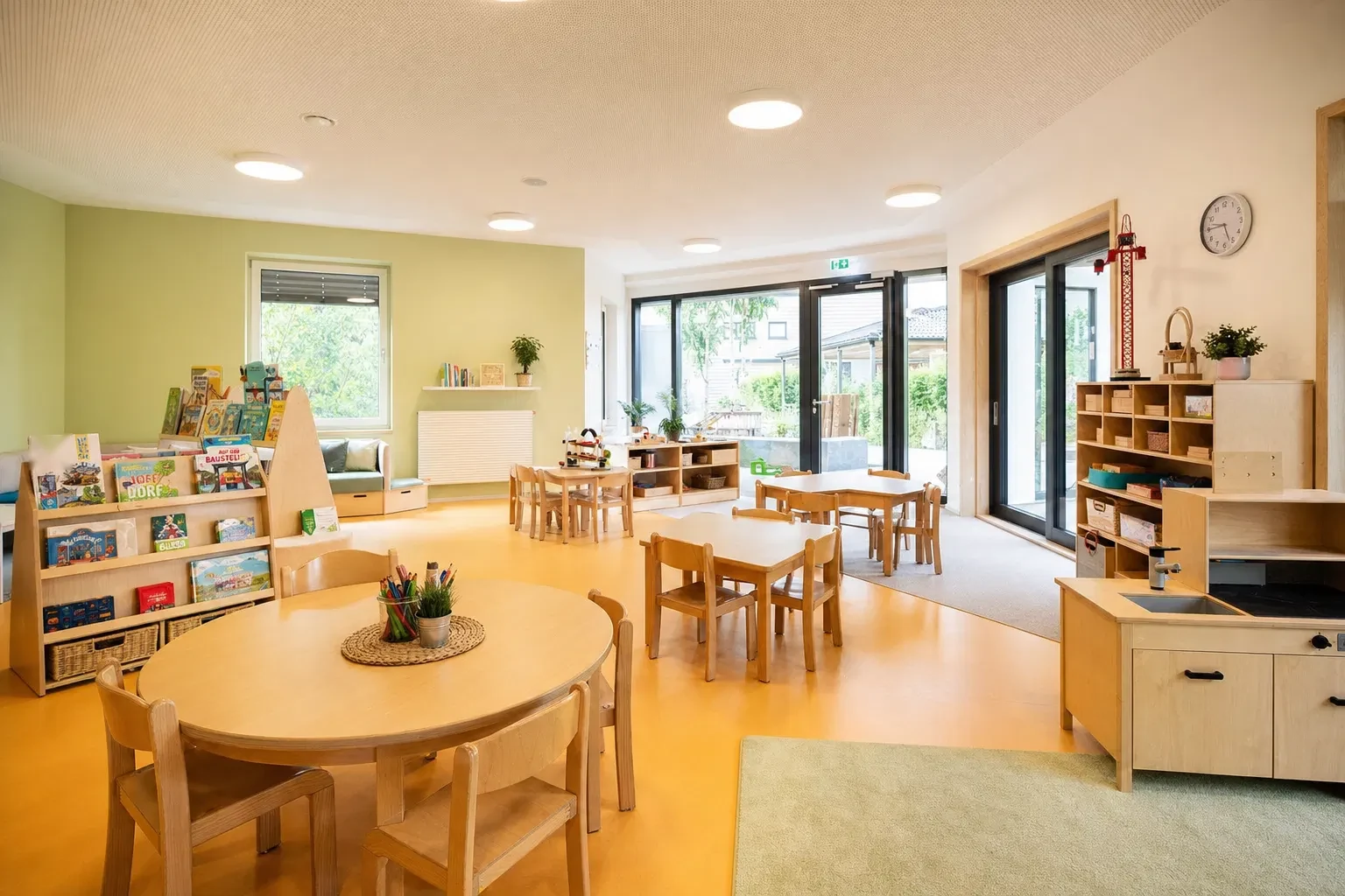 Heller Gruppenraum mit kleinen Tischen und Stühlen, Bücherregalen und großen Fenstertüren zum Garten