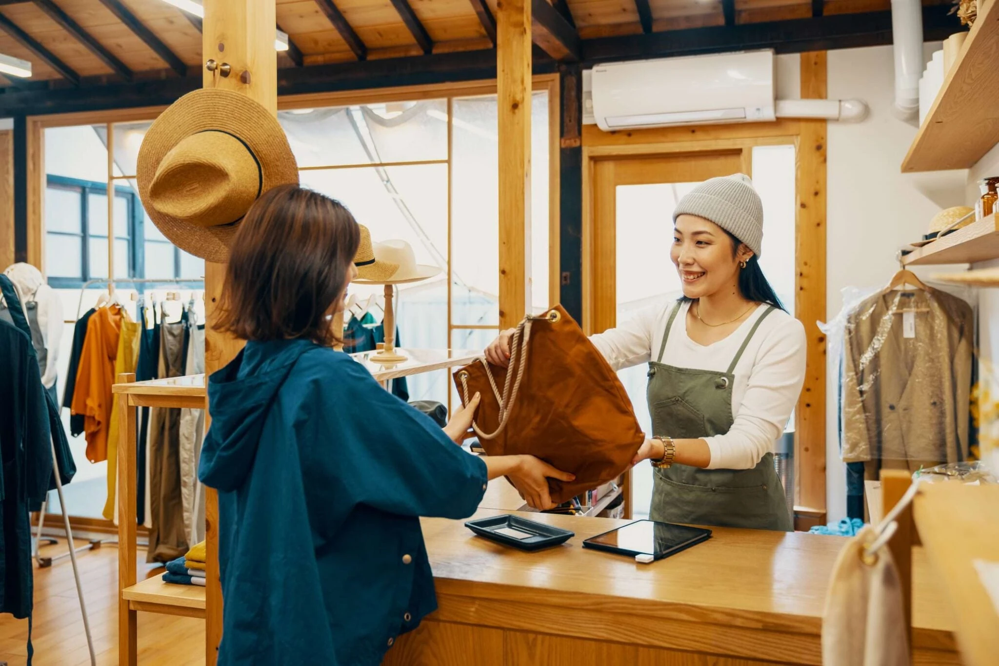 Inhaberin mit Schürze und Mütze reicht in ihrem hellen Bekleidungsladen einer Kundin eine Tasche über die Ladentheke