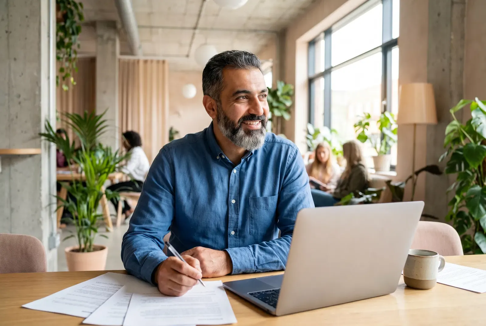 Lächelnder Gründer mit grauem Bart arbeitet an einem Laptop mit Unterlagen in einem hellen Coworking-Space