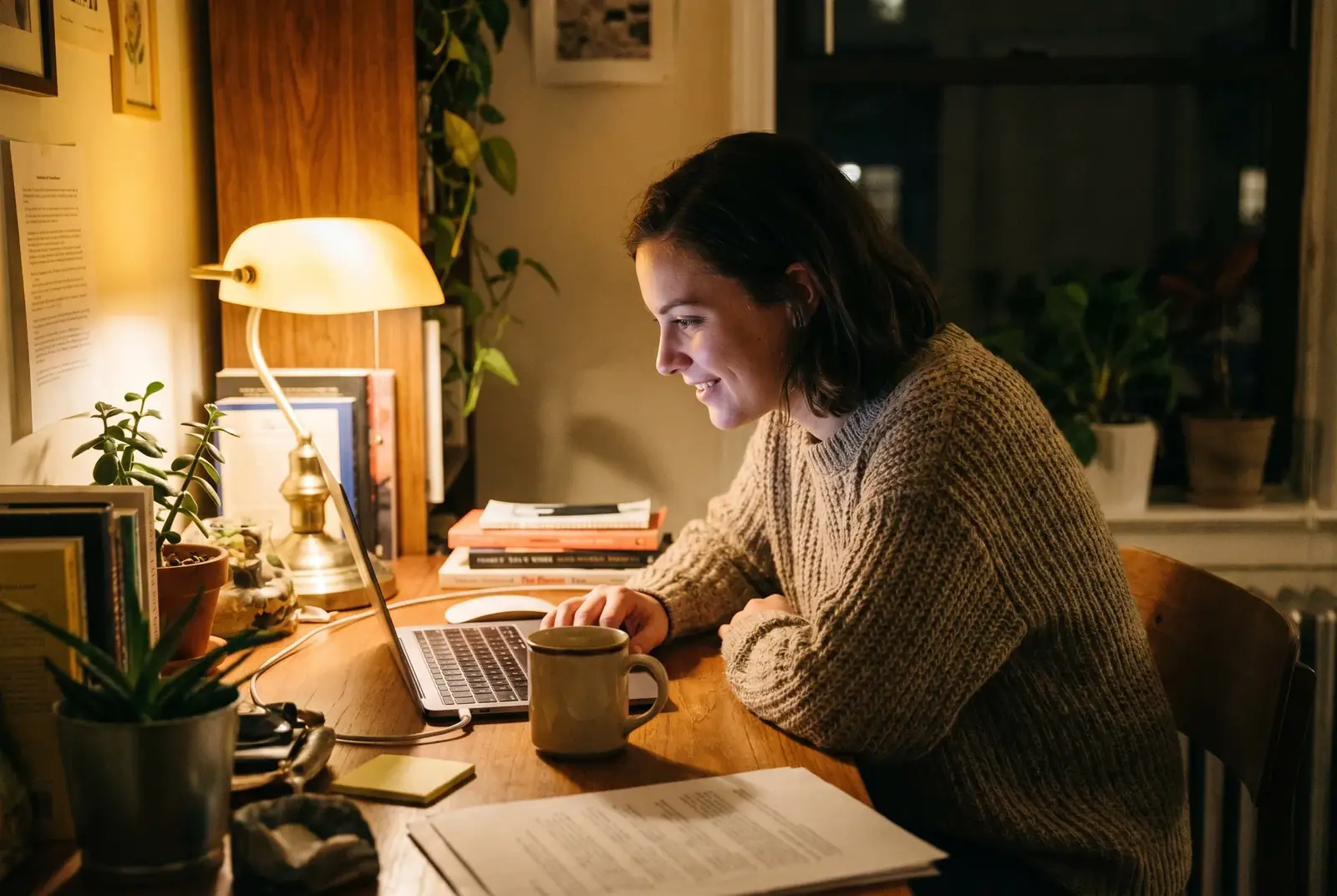 Lächelnde Frau arbeitet abends bei Lampenlicht am Laptop an ihrem Schreibtisch zu Hause