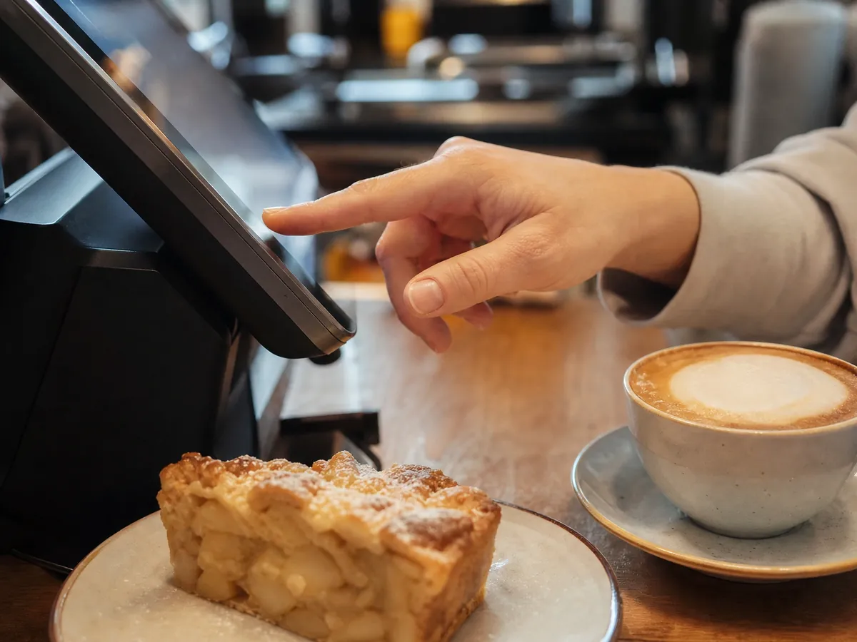 Hand bedient die Kasse am Tresen eines Cafés neben Kuchen und Cappuccino, Thema Kassenpflicht