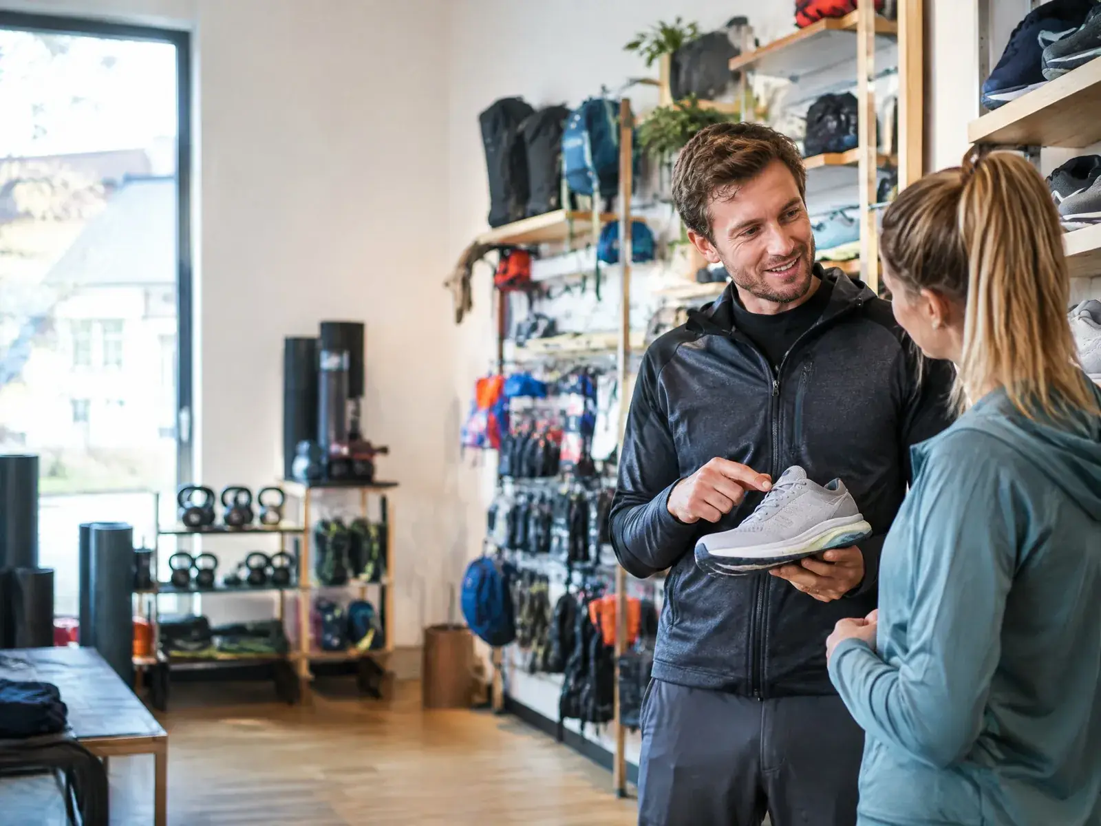 Verkäufer im Sportgeschäft zeigt einer Kundin einen Laufschuh, dahinter Regale mit Sportausrüstung
