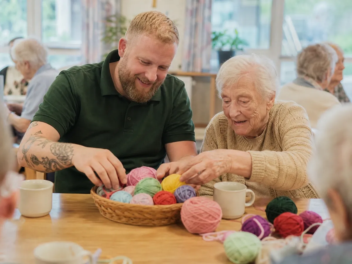 Betreuungskraft in einer Tagespflege beschäftigt sich am Gruppentisch mit einer Seniorin beim Basteln