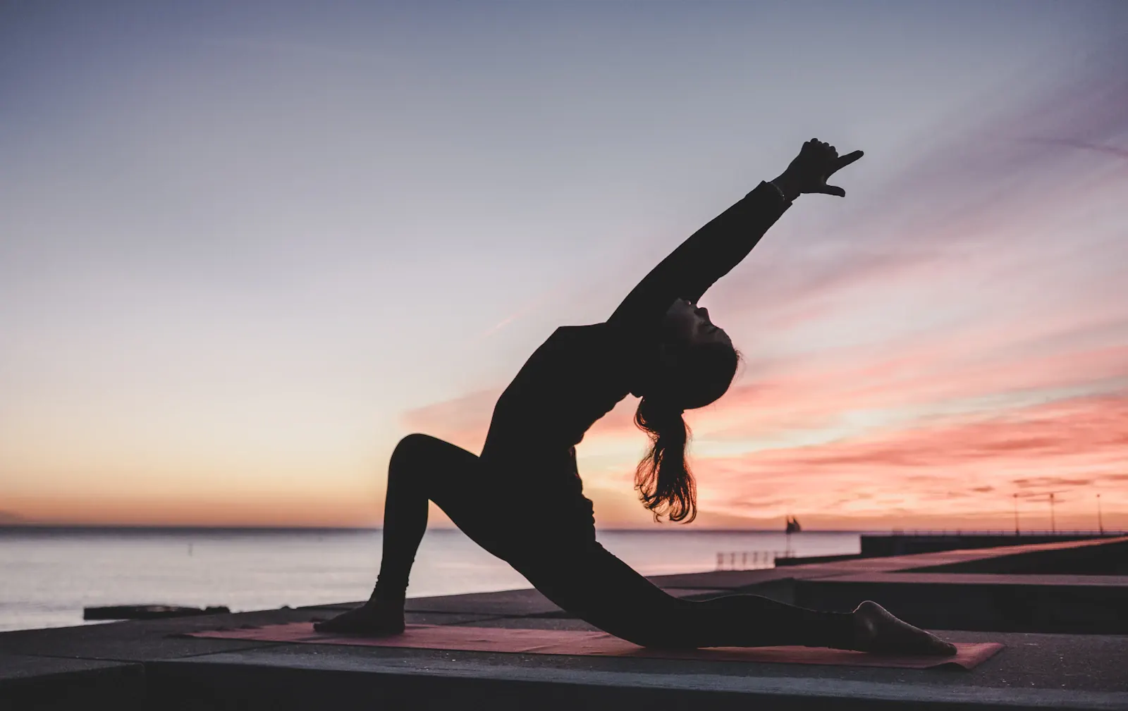 Yogalehrerin als Silhouette in einer Yogahaltung auf einer Matte am Wasser bei Sonnenaufgang