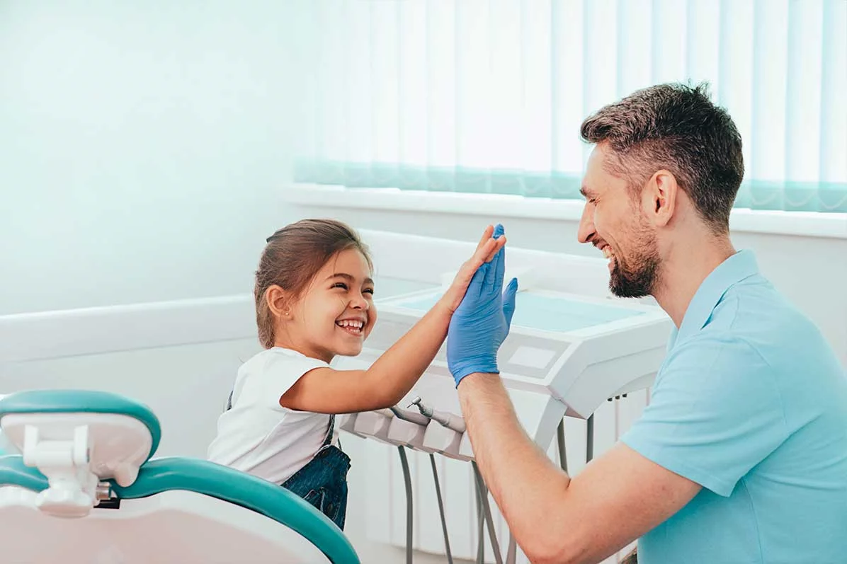 Zahnarzt in hellblauem Kasack mit blauen Handschuhen klatscht mit einem lachenden Mädchen auf dem Behandlungsstuhl ab